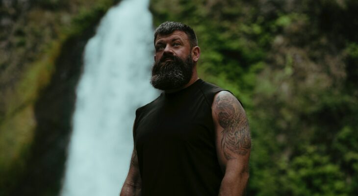 a man with a beard standing in front of a waterfall